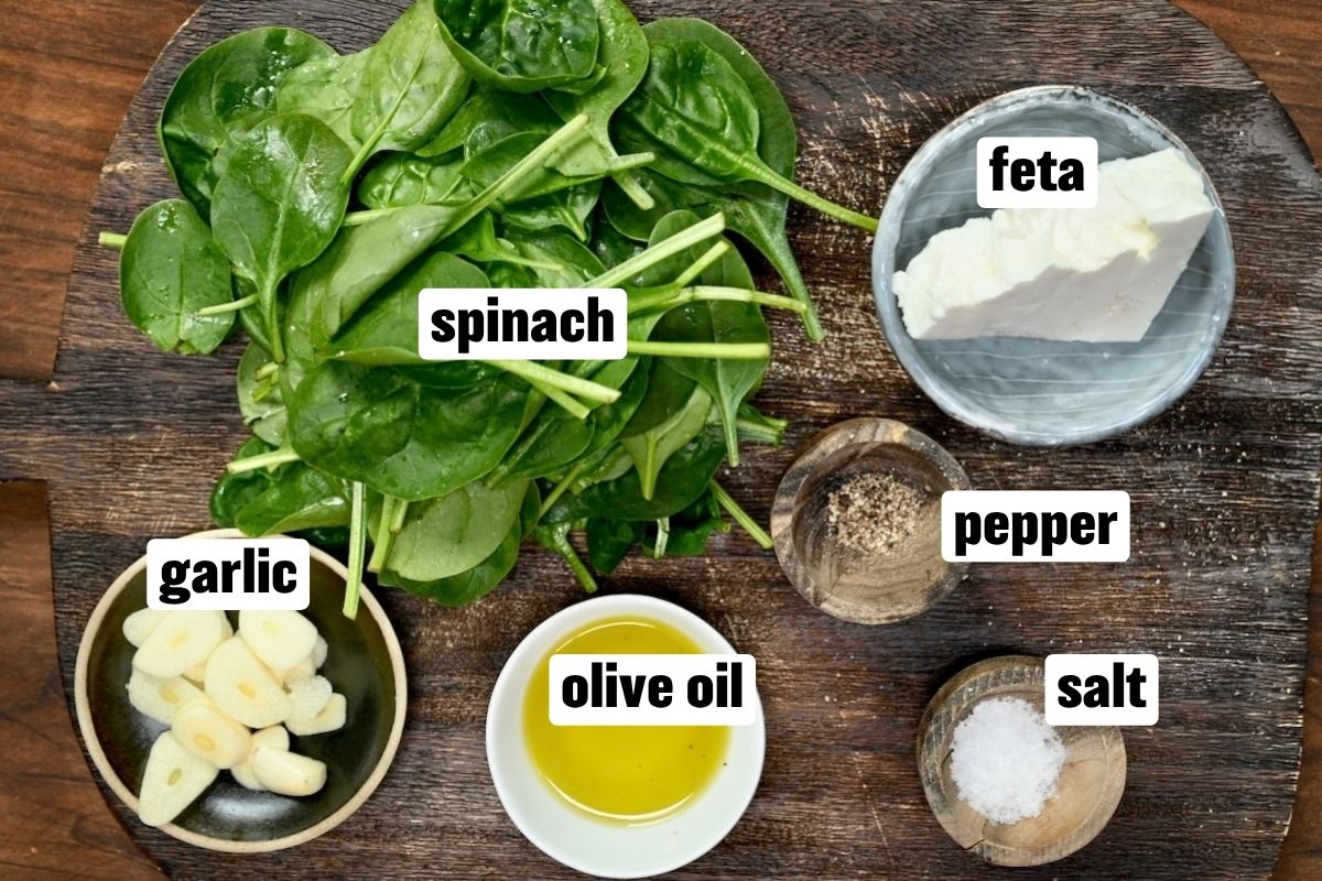 An overhead ingredient flat lay arranged on a dark wooden cutting board featuring fresh baby spinach leaves, peeled garlic cloves in a small dish, a wedge of feta cheese in a grey bowl, olive oil in a white ramekin, and small wooden pinch bowls containing sea salt and black pepper.