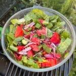 A top-down shot of a large glass mixing bowl packed with a vibrant garden salad made of chopped lettuce, chunks of ripe red tomatoes, cucumber slices, and slivered red onions, dusted with ground sumac and herbs.