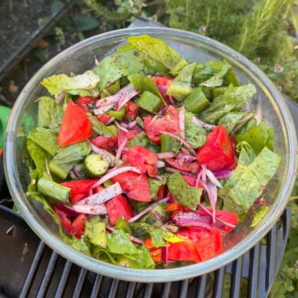 A top-down shot of a large glass mixing bowl packed with a vibrant garden salad made of chopped lettuce, chunks of ripe red tomatoes, cucumber slices, and slivered red onions, dusted with ground sumac and herbs.