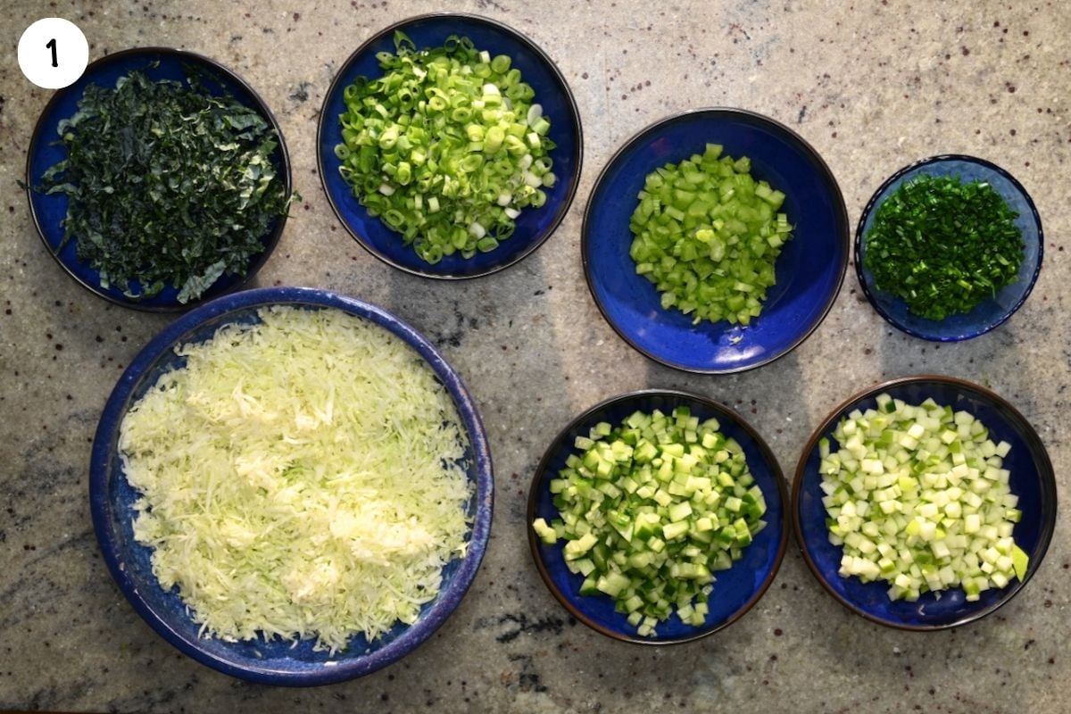 A top-down view of finely chopped salad ingredients separated into blue ceramic bowls, including shredded cabbage, chopped kale, sliced scallions, diced celery, minced chives, diced cucumbers, and diced green apples.