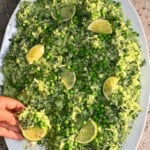 A close-up view of Green Goddess salad spread over a platter, loaded with green peas, fresh dill, and lime slices, with a hand holding a chip topped with salad and a slice of lime.