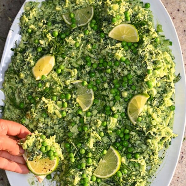A close-up view of Green Goddess salad spread over a platter, loaded with green peas, fresh dill, and lime slices, with a hand holding a chip topped with salad and a slice of lime.