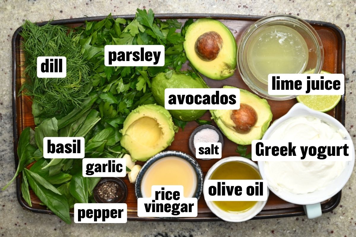 A top-down view of fresh dressing ingredients on a wooden tray, including dill, parsley, basil, garlic, halved avocados, lime juice, Greek yogurt, olive oil, rice vinegar, salt, and pepper.