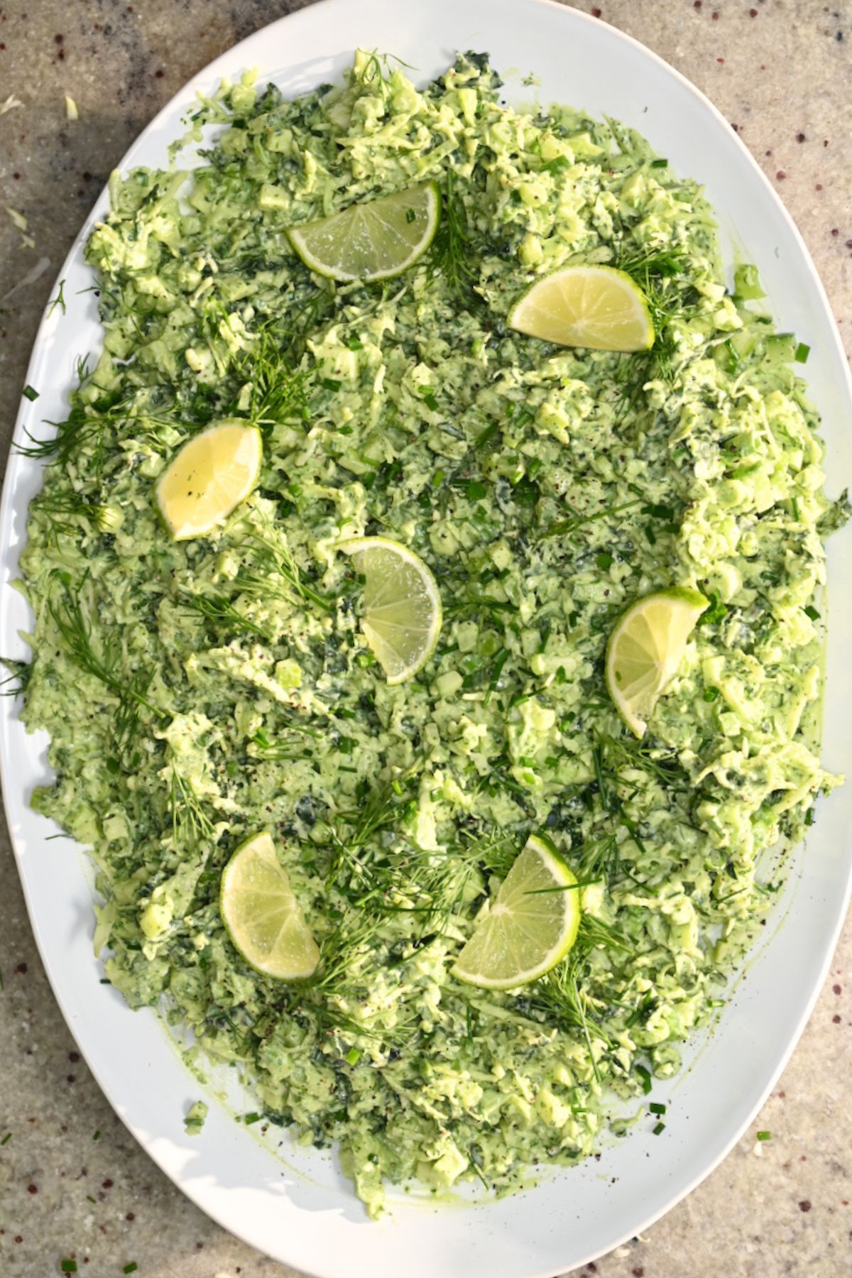An overhead view of creamy Green Goddess salad spread evenly across a white oval platter, garnished with fresh lime wedges, sprigs of dill, and chopped chives.