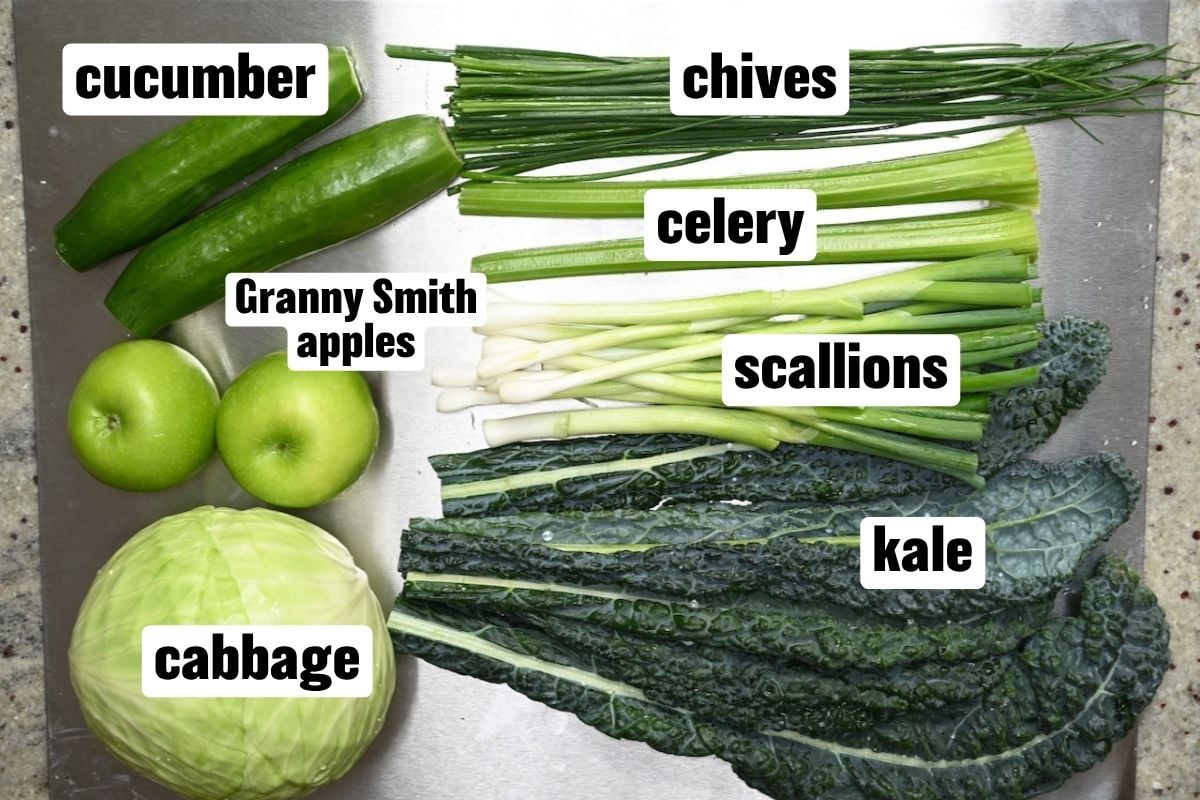 A top-down view of fresh green ingredients including cucumbers, chives, celery, scallions, Granny Smith apples, cabbage, and kale arranged on a cutting board.