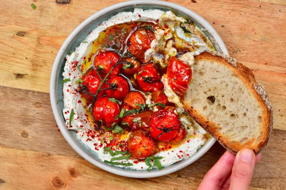 A shallow ceramic bowl filled with creamy labneh, topped with cherry tomato confit on the vine, warm olive oil, chili flakes, and fresh herbs, served with crusty bread dipping into the dish.
