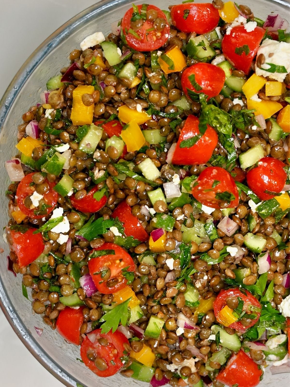 A detailed close-up shot of a glass bowl filled with vibrant lentil salad, showing cooked green lentils, juicy cherry tomatoes, diced cucumbers, yellow bell peppers, red onion, and crumbled feta cheese.
