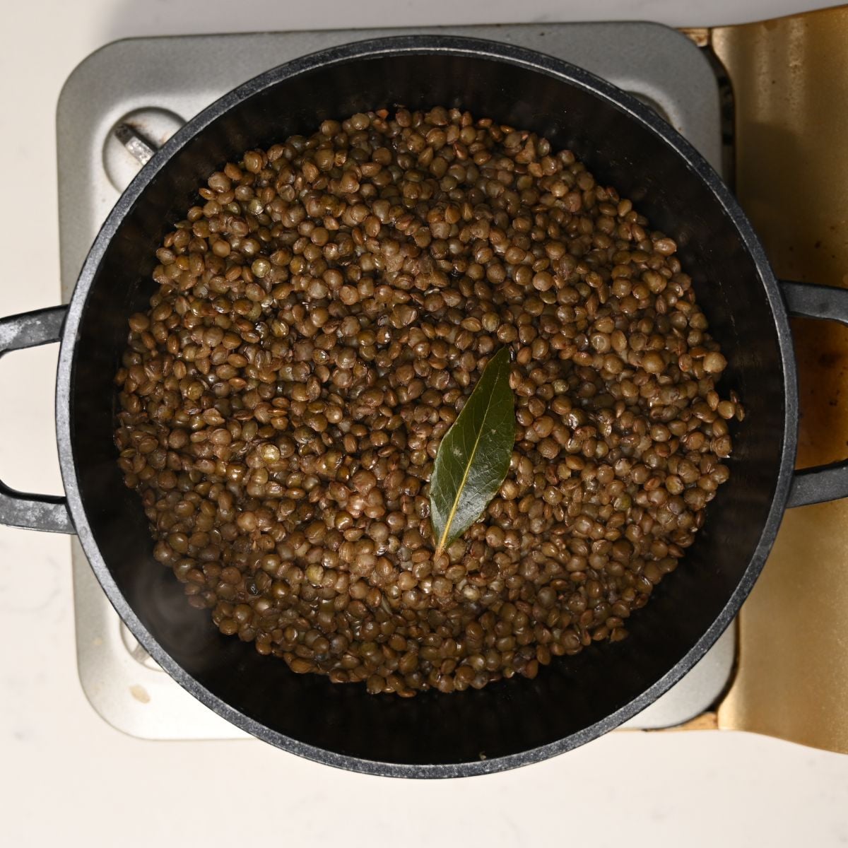 A black cooking pot filled with cooked green lentils simmered with a dried bay leaf on top.