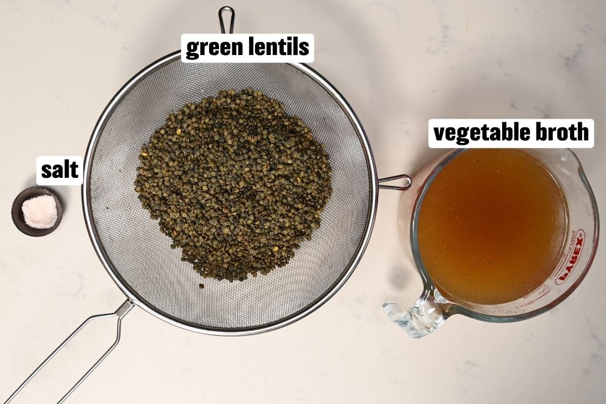 An ingredient setup featuring green lentils inside a fine-mesh strainer, a small pinch bowl of salt, and a glass measuring cup filled with vegetable broth.