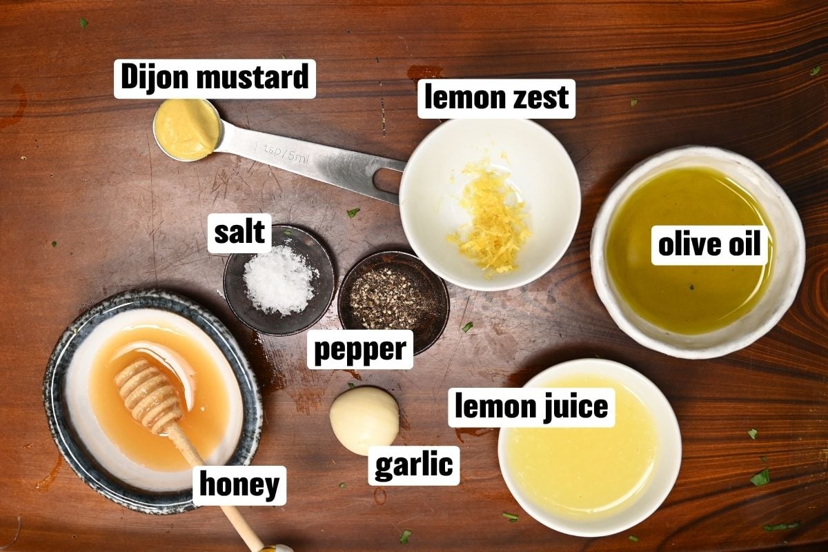 An ingredient flat lay on a wooden tray displaying small bowls of honey, lemon zest, lemon juice, olive oil, garlic, salt, pepper, and a measuring spoon with Dijon mustard.