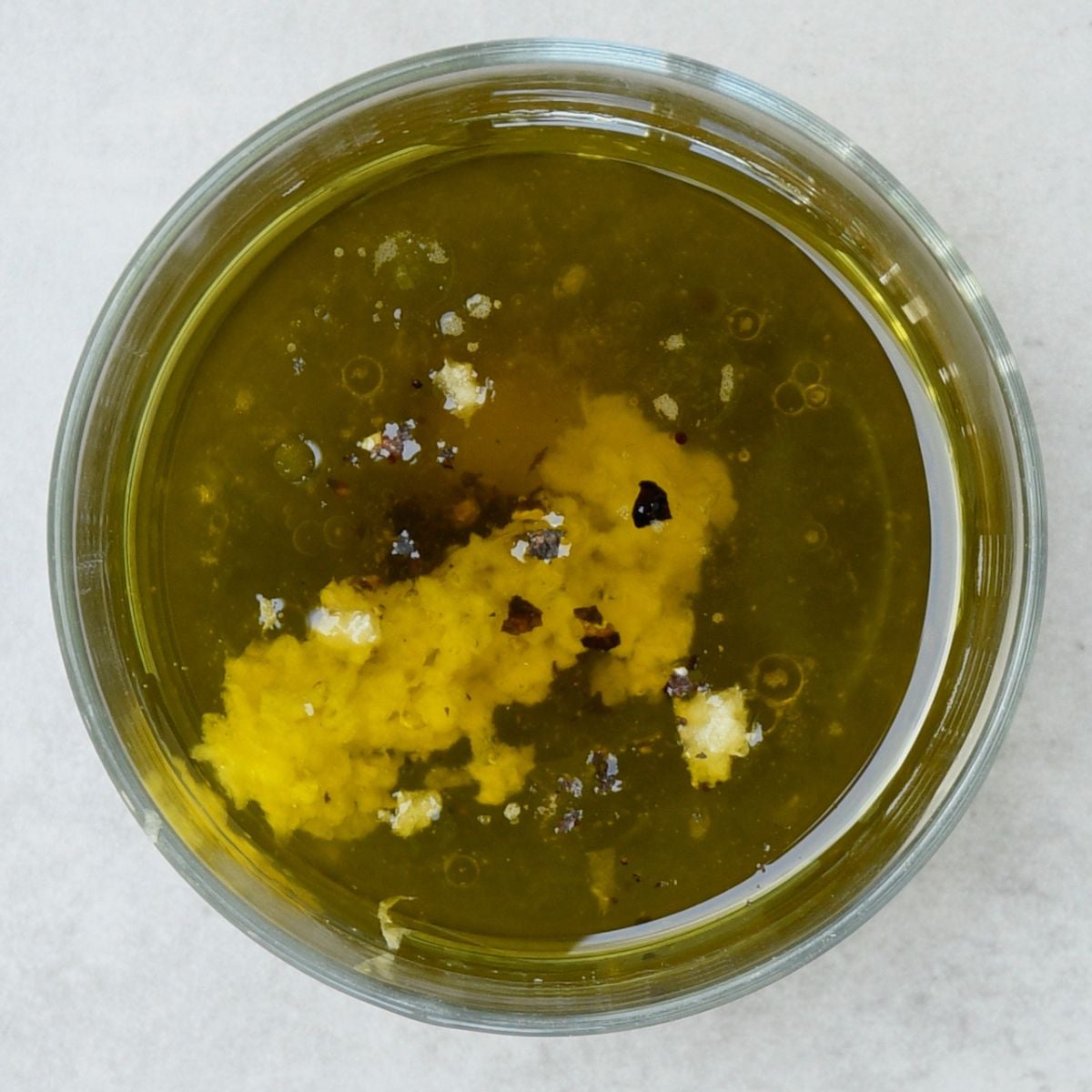 A top-down view of olive oil, lemon zest, Dijon mustard, and seasonings unmixed inside a glass bowl.