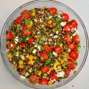 A top-down view of a clear glass bowl filled with cooked green lentils tossed together with halved cherry tomatoes, diced cucumbers, yellow bell peppers, red onion, parsley, and crumbled feta cheese.