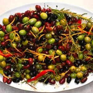 A close-up view of mixed green and black marinated olives tossed with orange and lemon zest strips, fresh rosemary sprigs, thyme, and red chili peppers.