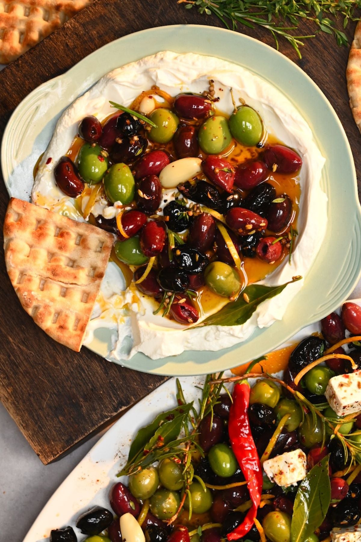 A overhead view of a light green plate filled with creamy labneh topped with warm marinated olives, oil, and spices, served with pita chips.