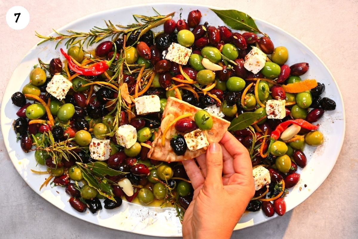A top-down view of an oval platter filled with mixed marinated olives, feta cheese cubes, fresh herbs, and red chilies, with a hand holding a piece of pita bread topped with an olive and feta.