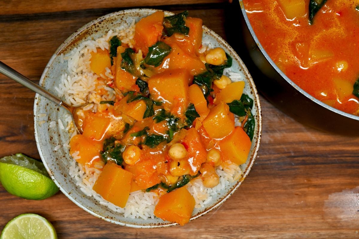 A top-down view of a ceramic bowl filled with white rice and topped with pumpkin chickpea curry next to lime wedges on a wooden table.