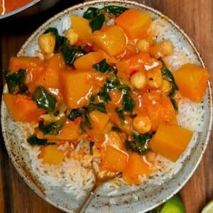 A close-up top-down view of a bowl filled with white rice and topped with rich pumpkin chickpea curry with spinach and a spoon sticking out.