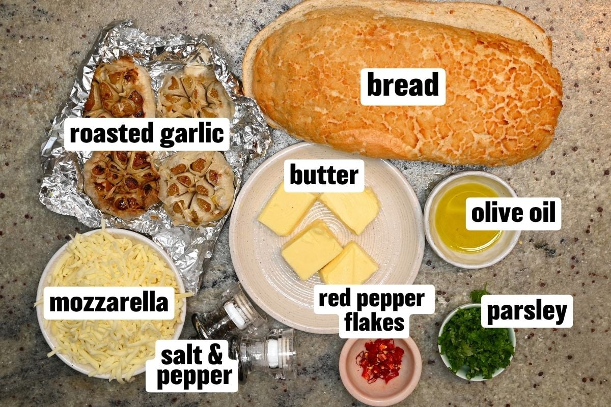 A flat-lay view of roasted garlic bread ingredients, including a loaf of bread, roasted garlic bulbs wrapped in foil, butter, shredded mozzarella, olive oil, parsley, red pepper flakes, salt, and pepper arranged on a countertop.