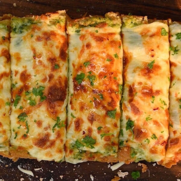A top-down close-up view of freshly baked, cheesy roasted garlic bread slices topped with herbs on a wooden cutting board.