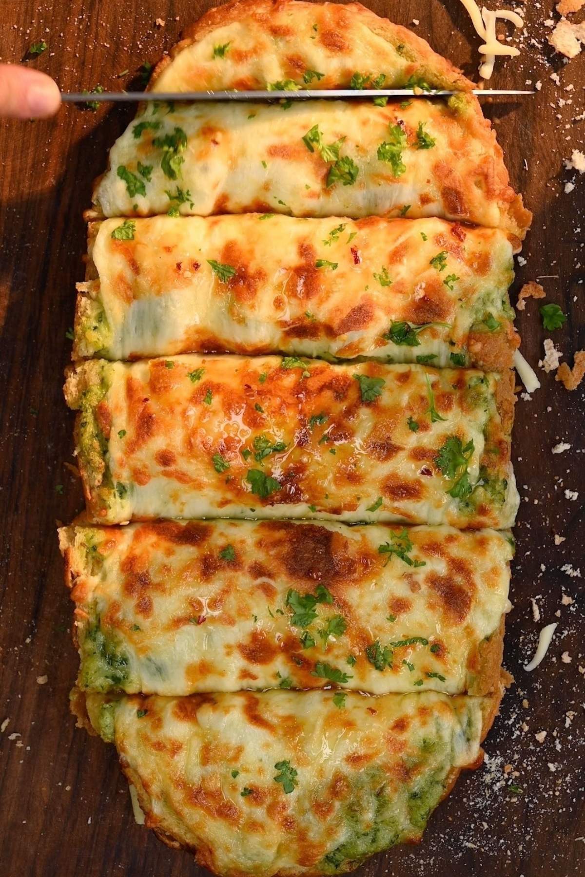 A top-down view of a baked, cheesy roasted garlic bread loaf being sliced into individual portions with a knife on a wooden cutting board.