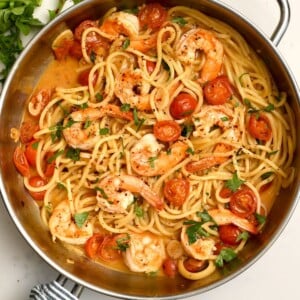 A detailed overhead view of finished shrimp pasta in a stainless steel skillet, coated in a light sauce, garnished with parsley, sliced garlic, and crushed red pepper flakes.