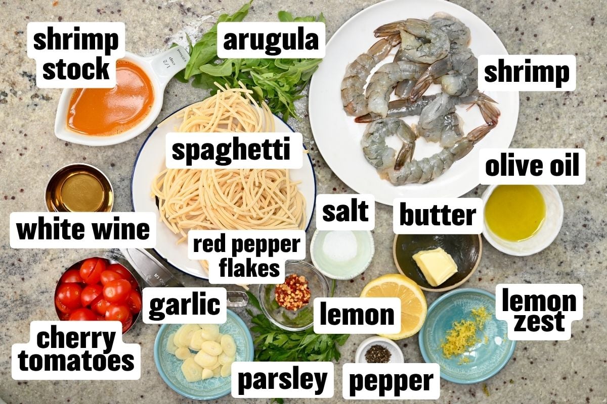 A top-down view of raw ingredients for shrimp pasta with text labels, including fresh shrimp, spaghetti, cherry tomatoes, garlic, white wine, shrimp stock, arugula, lemon, lemon zest, butter, olive oil, parsley, red pepper flakes, salt, and pepper.