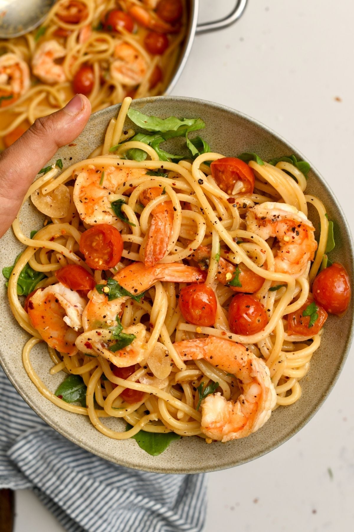 A hand holding a grey ceramic bowl filled with shrimp pasta, cherry tomatoes, fresh arugula, black pepper, and chili flakes, with the cooking skillet visible in the background.