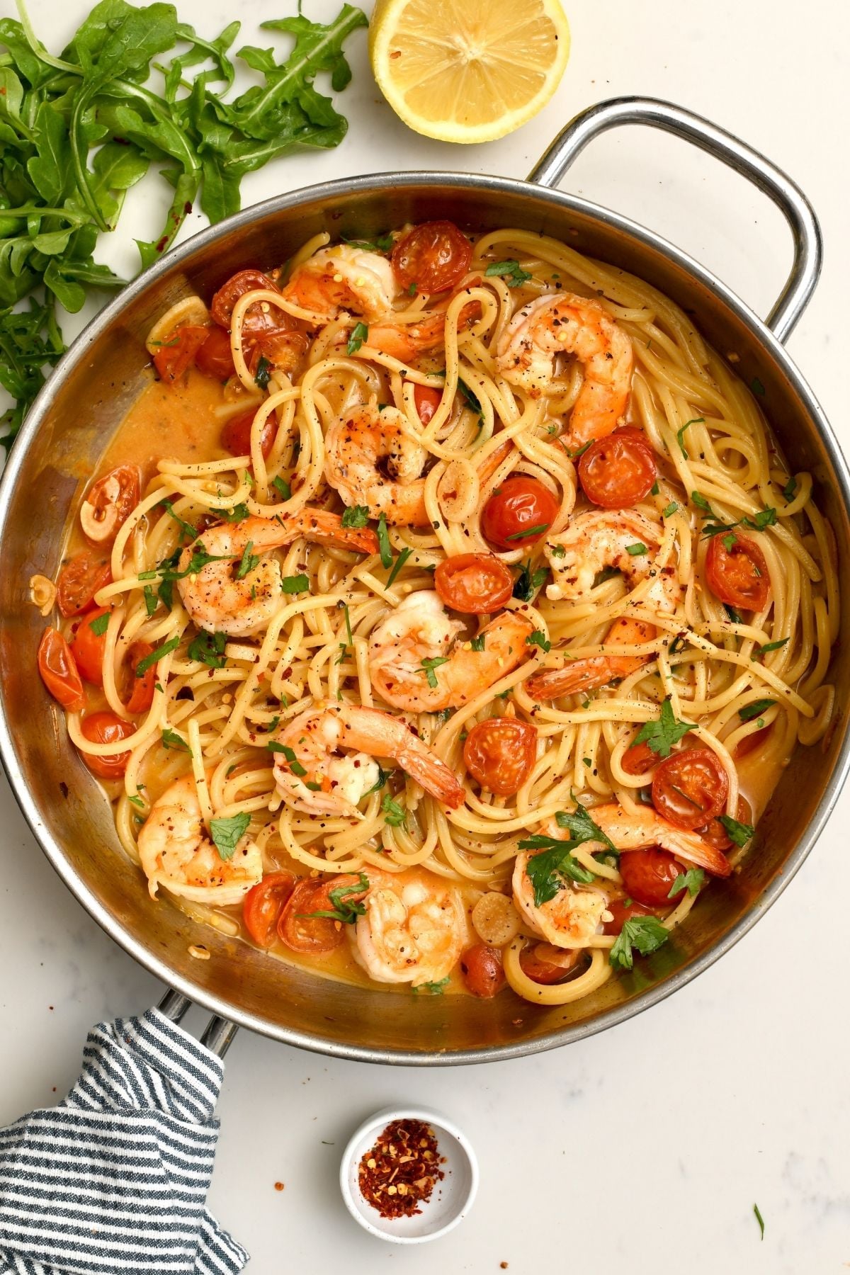 A top-down view of a stainless steel skillet filled with fresh shrimp pasta tossed with cherry tomatoes, parsley, and sauce, garnished with fresh arugula and a halved lemon on the side.