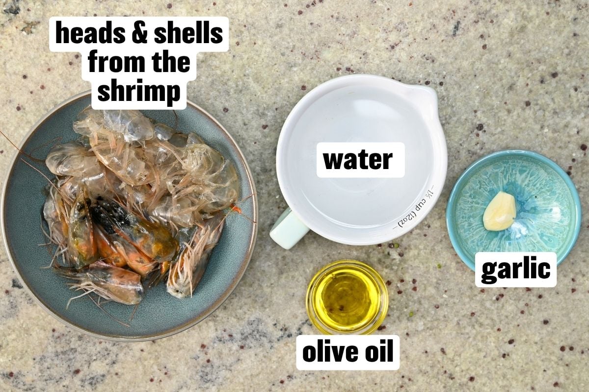 A top-down view of ingredients for making homemade shrimp stock, with text labels pointing to shrimp heads and shells in a bowl, a measuring cup of water, olive oil, and garlic.
