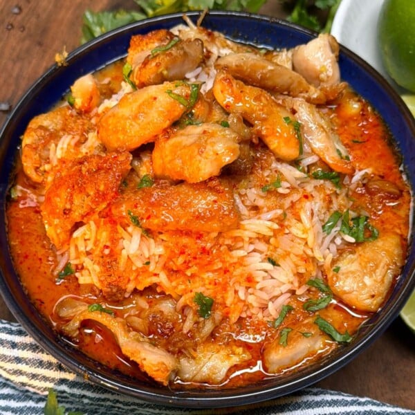 A top-down view of a dark blue serving bowl filled with jasmine rice and rich red curry broth, topped with tender seared chicken pieces and garnished with fresh cilantro, with lime wedges on the side.