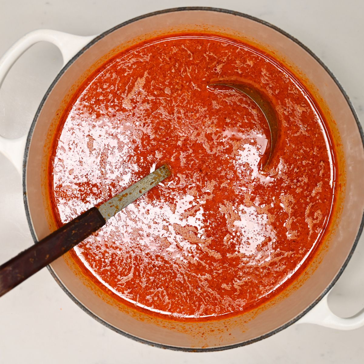 A top-down view of vibrant red Thai curry broth simmering inside a white Dutch oven pot with a wooden ladle resting in the liquid.