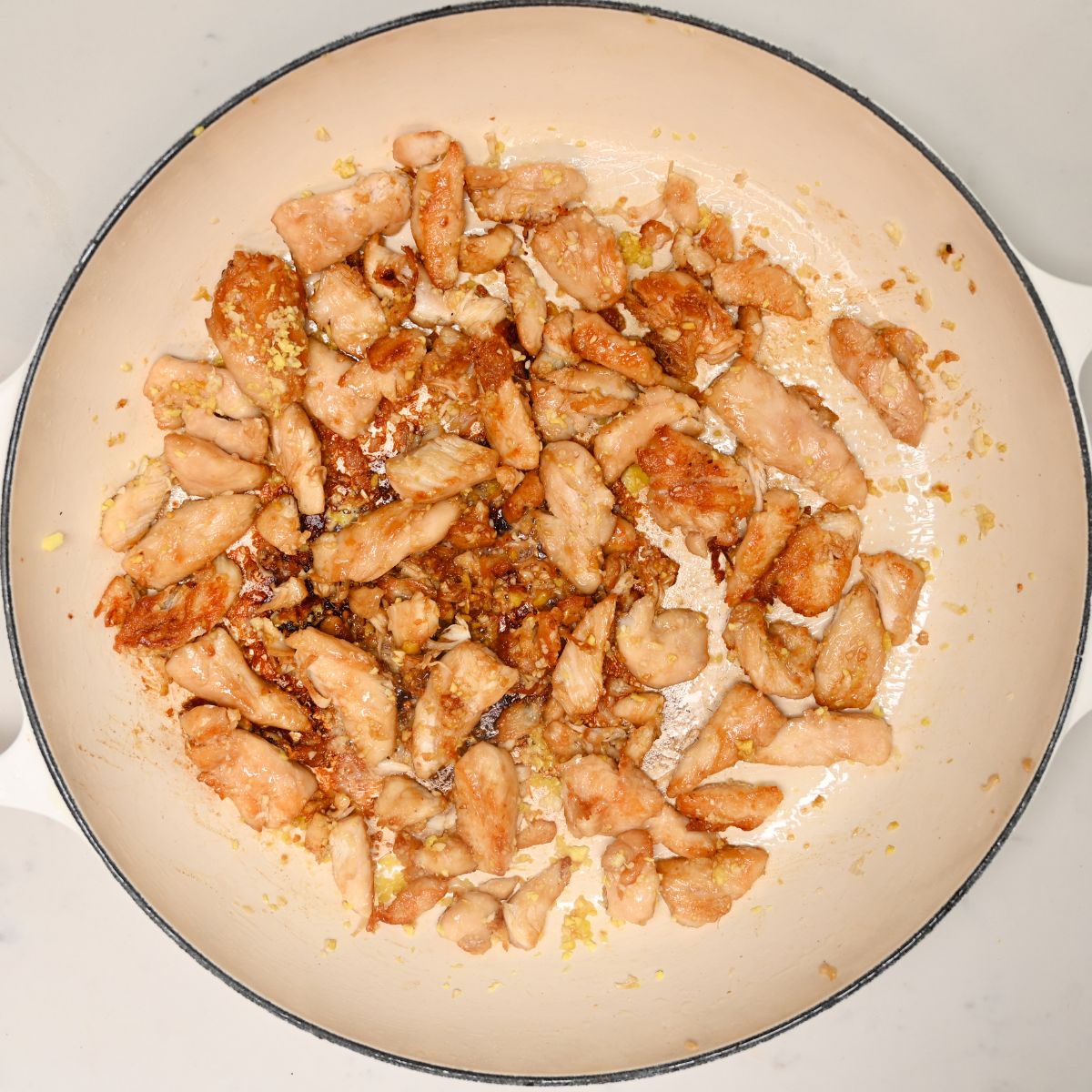A top-down view of bite-sized chicken pieces searing to a light golden brown with garlic and ginger inside a large enamel skillet.