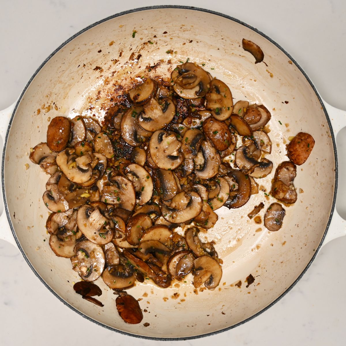 A top-down view of sliced brown mushrooms sautéing until tender with herbs and aromatics in a large light-colored skillet.