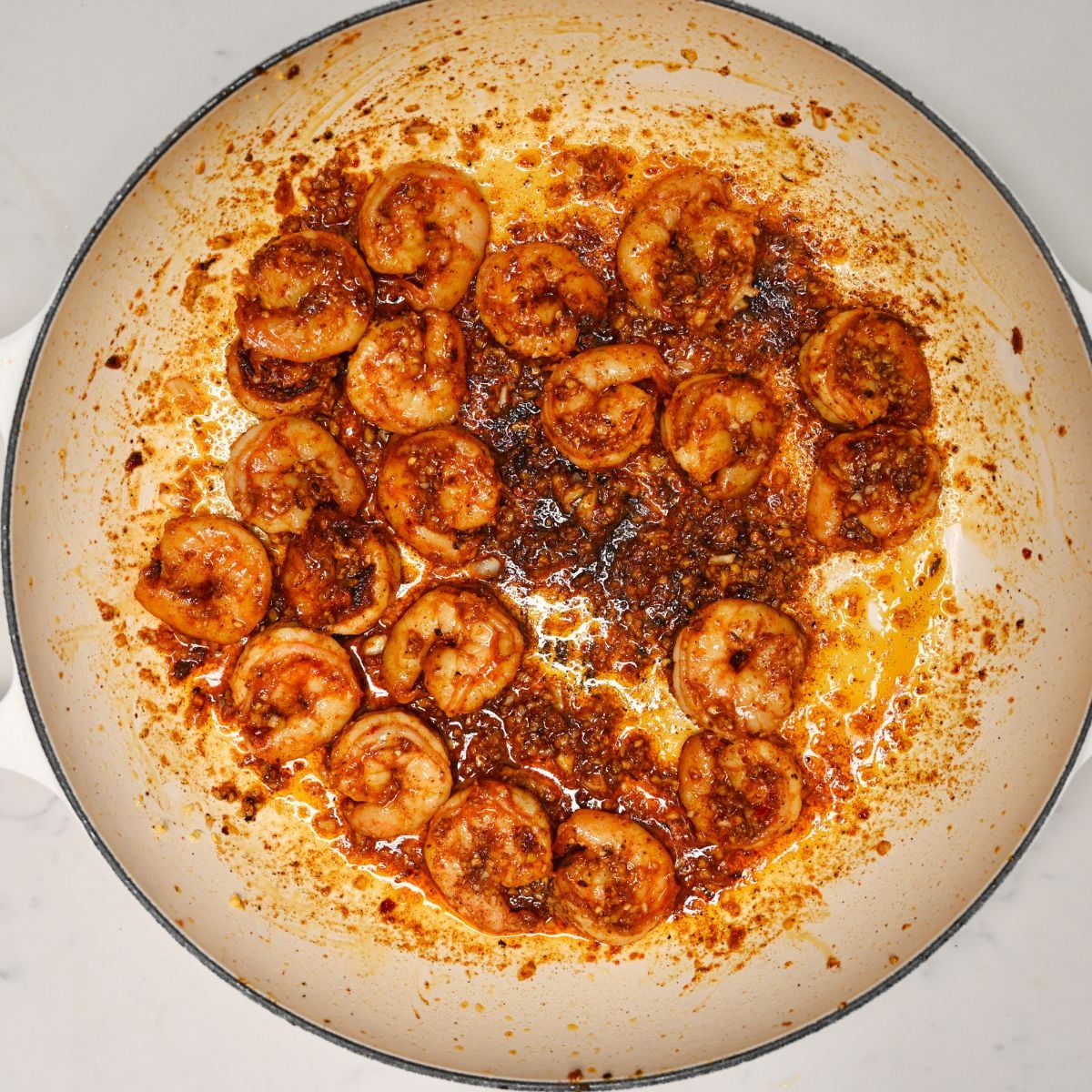 A top-down view of seasoned prawns searing in a light-colored enamel skillet with garlic, ginger, and pan juices.