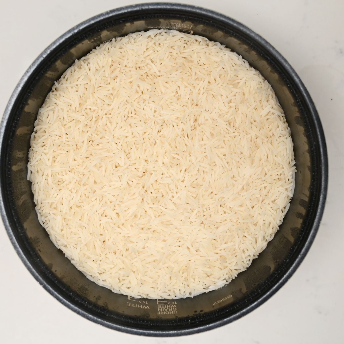 A top-down view of uncooked long-grain white rice spread evenly inside the inner pot of a rice cooker.