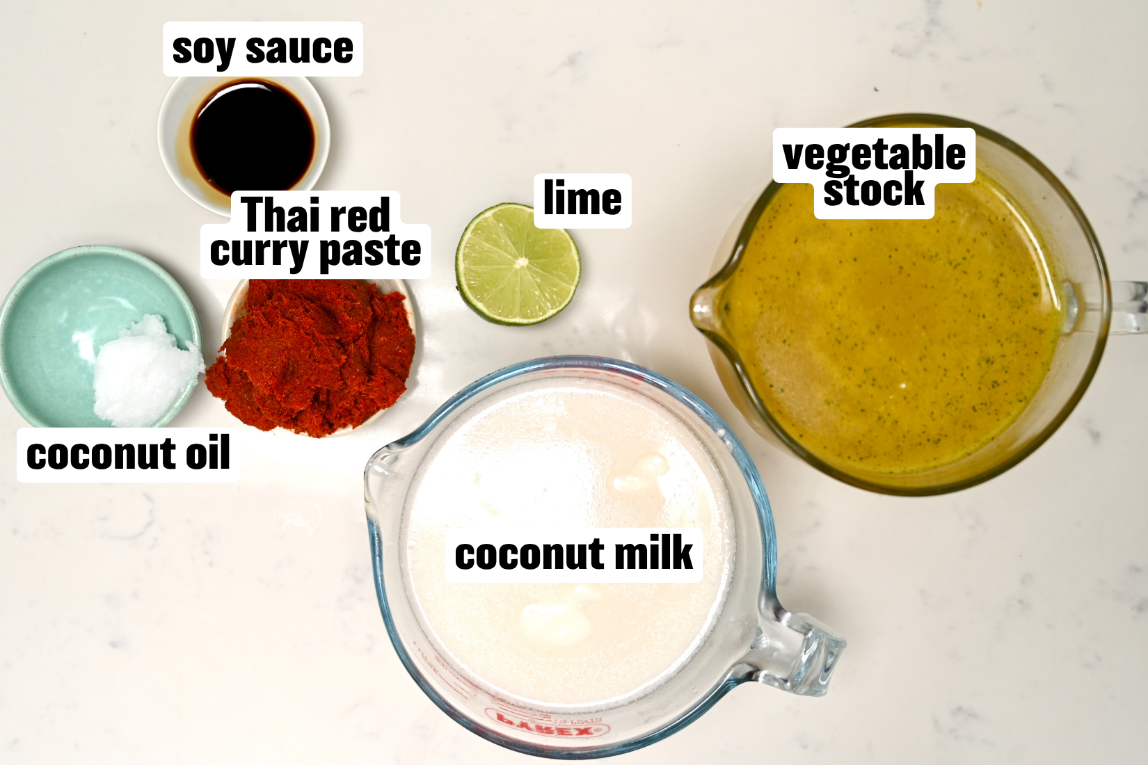 An ingredient flat lay arranged on a white surface featuring coconut milk, vegetable stock, Thai red curry paste, soy sauce, coconut oil, and a lime half.