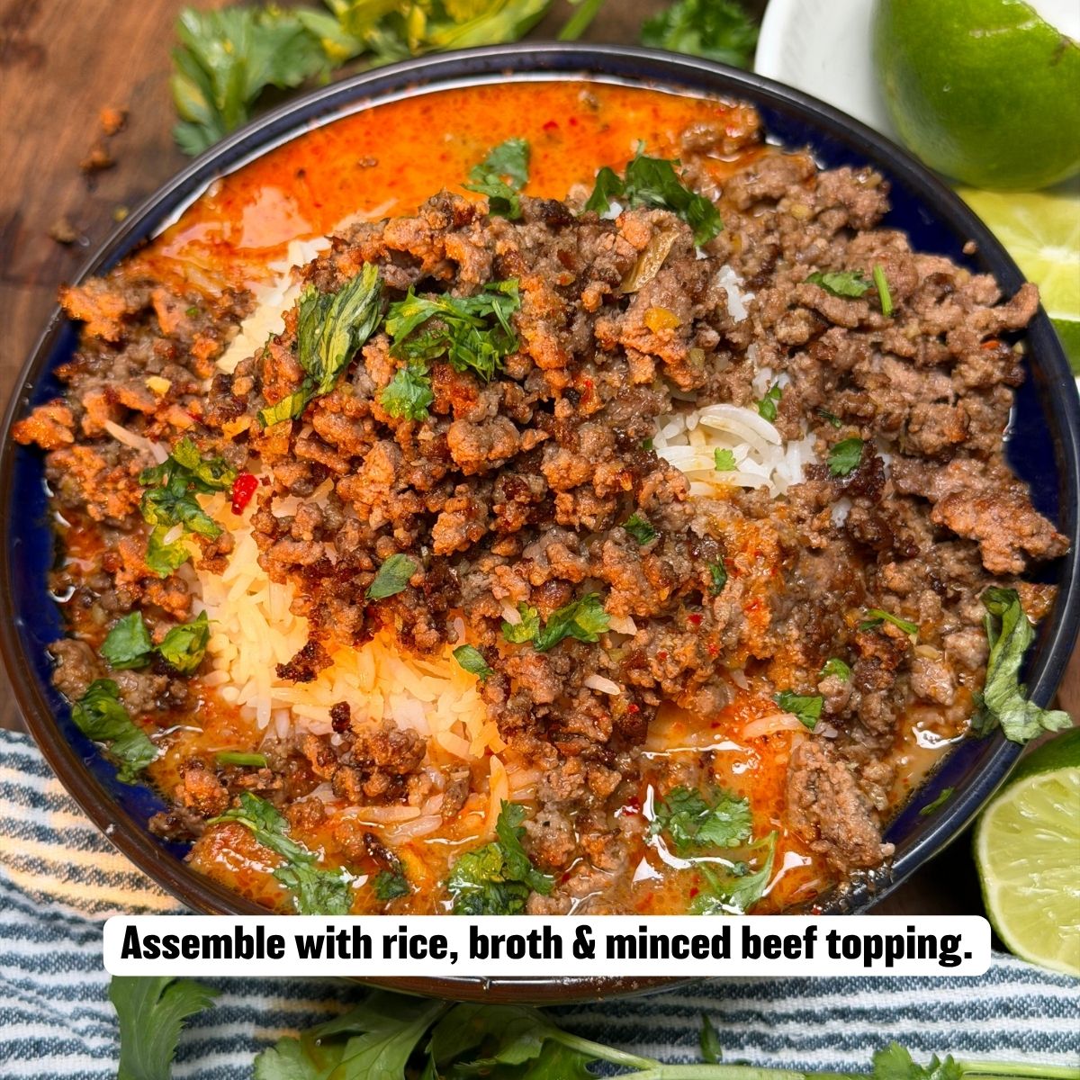 A top-down view of a dark blue serving bowl filled with rice and red curry broth, topped with seasoned browned minced beef and garnished with chopped fresh cilantro, served with lime halves on the side.