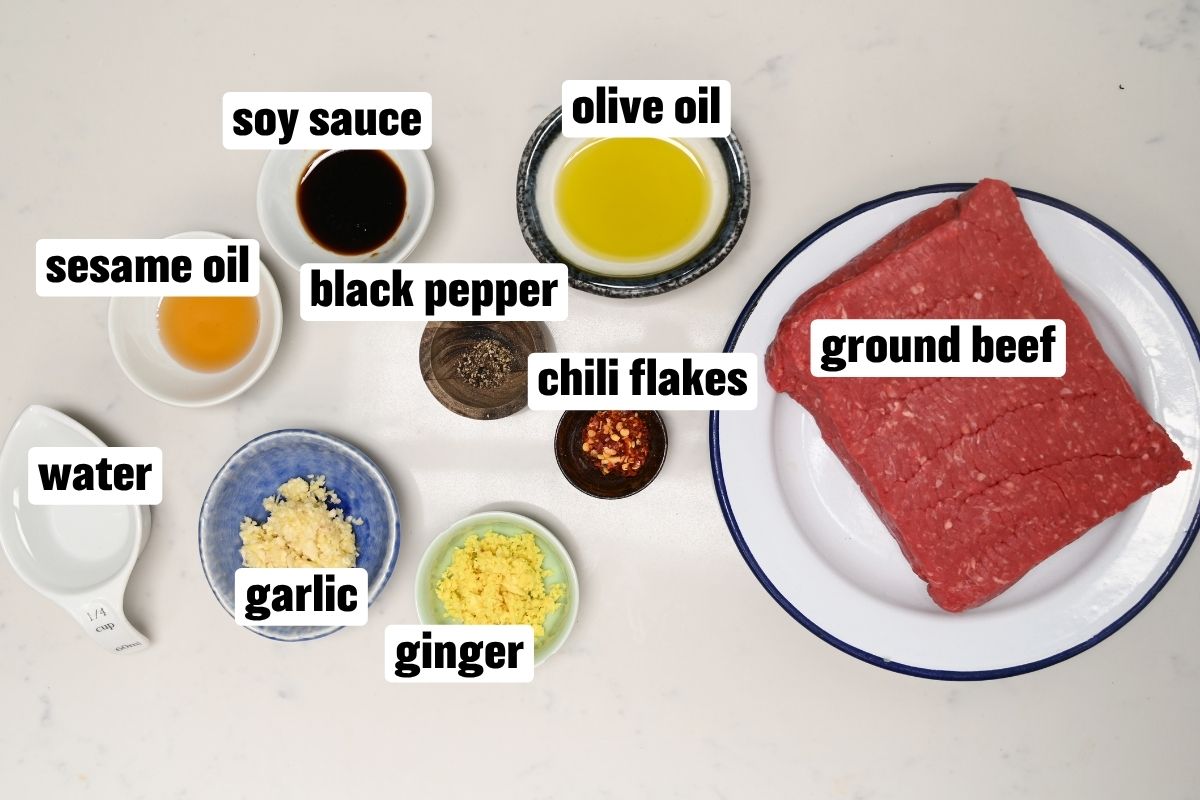 An ingredient flat lay arranged on a white surface featuring a white plate with fresh ground beef alongside small bowls and measuring cups filled with soy sauce, olive oil, sesame oil, water, minced garlic, grated ginger, chili flakes, and black pepper.