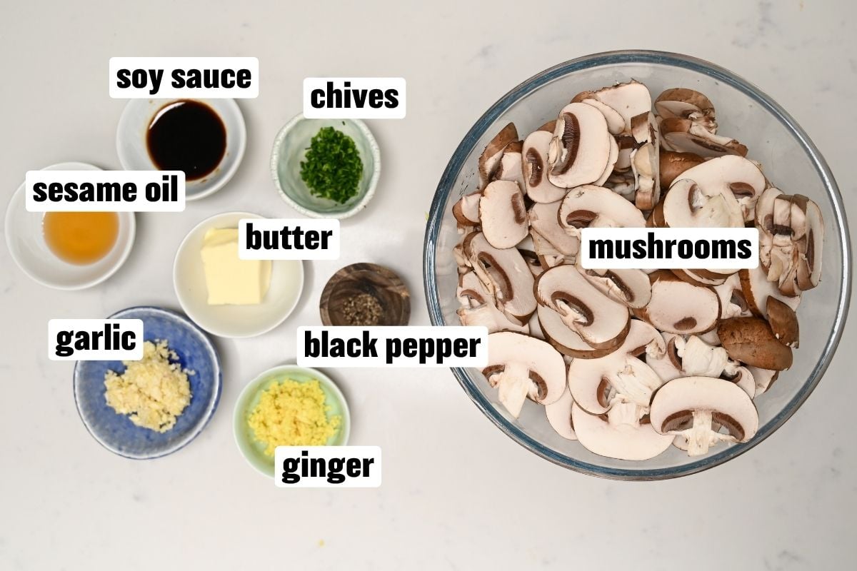 An ingredient flat lay arranged on a white surface featuring a large glass bowl of sliced brown mushrooms surrounded by small bowls containing soy sauce, sesame oil, butter, minced garlic, grated ginger, chopped chives, and black pepper.