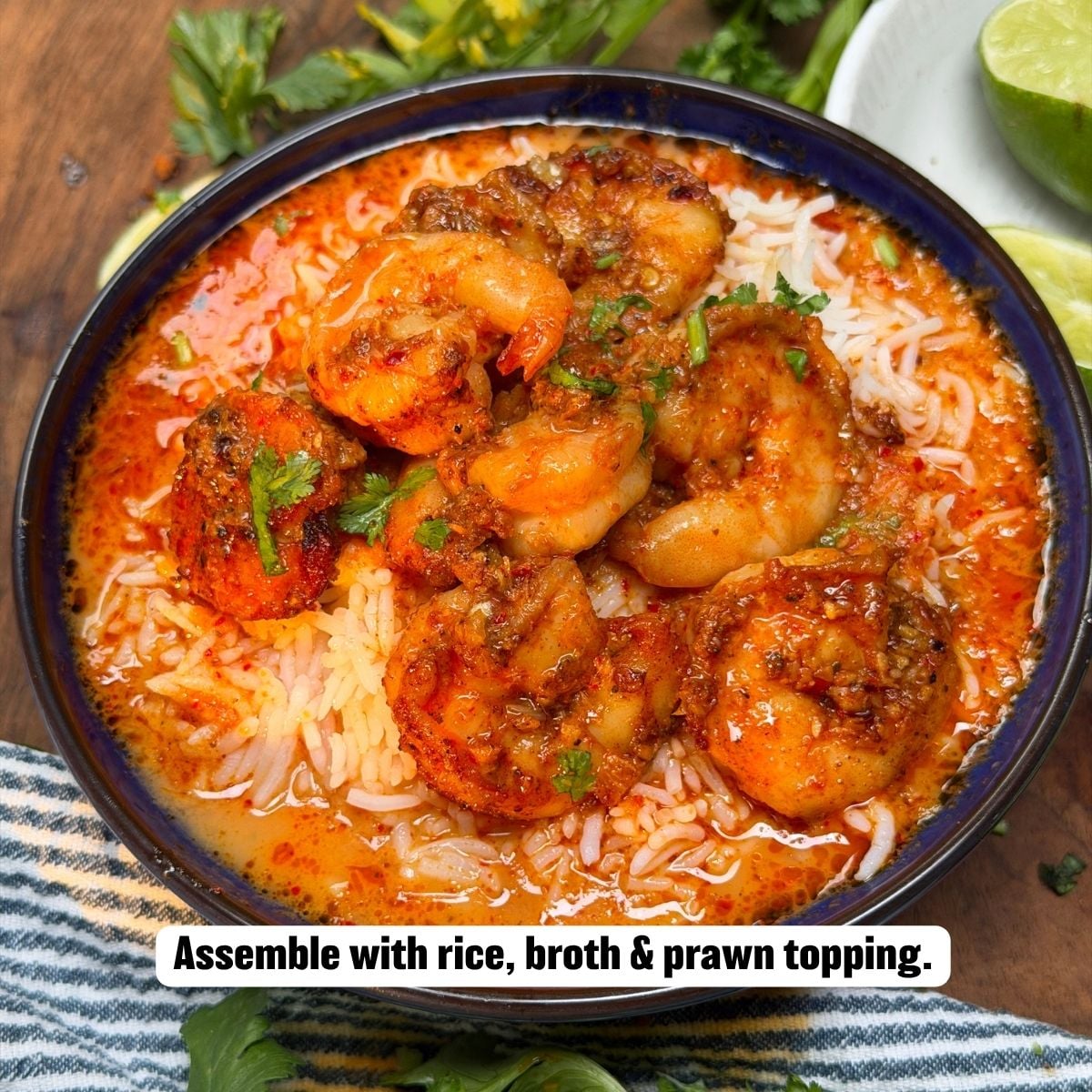 A top-down view of a dark serving bowl filled with rice and red curry broth, topped with seared, seasoned prawns and garnished with fresh cilantro, served with lime wedges on the side.