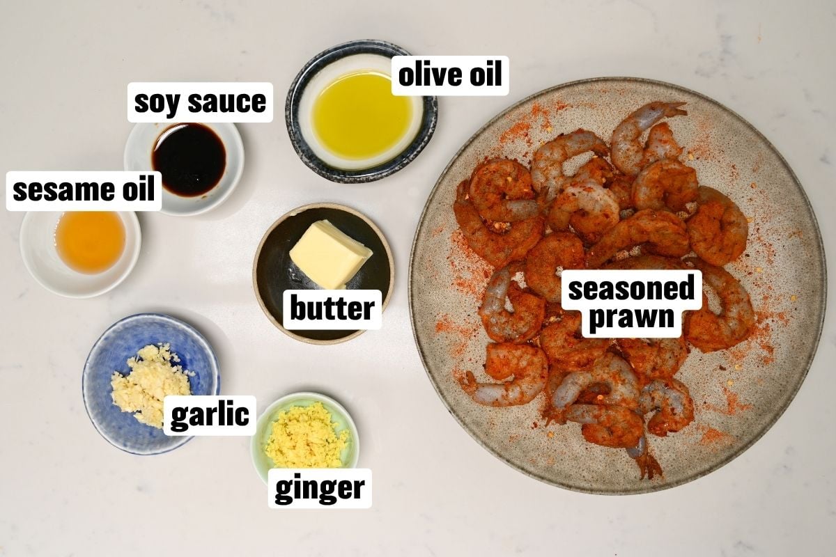 An ingredient flat lay arranged on a white surface featuring a large round plate filled with raw seasoned prawns alongside small bowls containing olive oil, butter, soy sauce, sesame oil, minced garlic, and grated ginger.