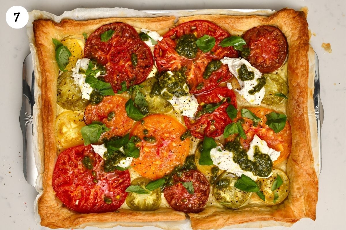 A top-down view of a baked heirloom tomato tart on a parchment-lined baking sheet with a golden-brown puff pastry crust, garnished with dollops of ricotta, basil pesto, and fresh basil leaves.