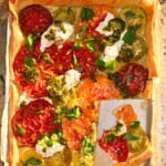 A top-down view of a baked heirloom tomato tart on a parchment-lined baking tray, topped with pesto, fresh cheese, and basil, with one slice cut out.