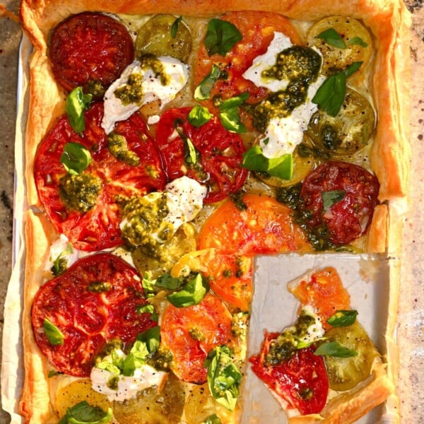 A top-down view of a baked heirloom tomato tart on a parchment-lined baking tray, topped with pesto, fresh cheese, and basil, with one slice cut out.