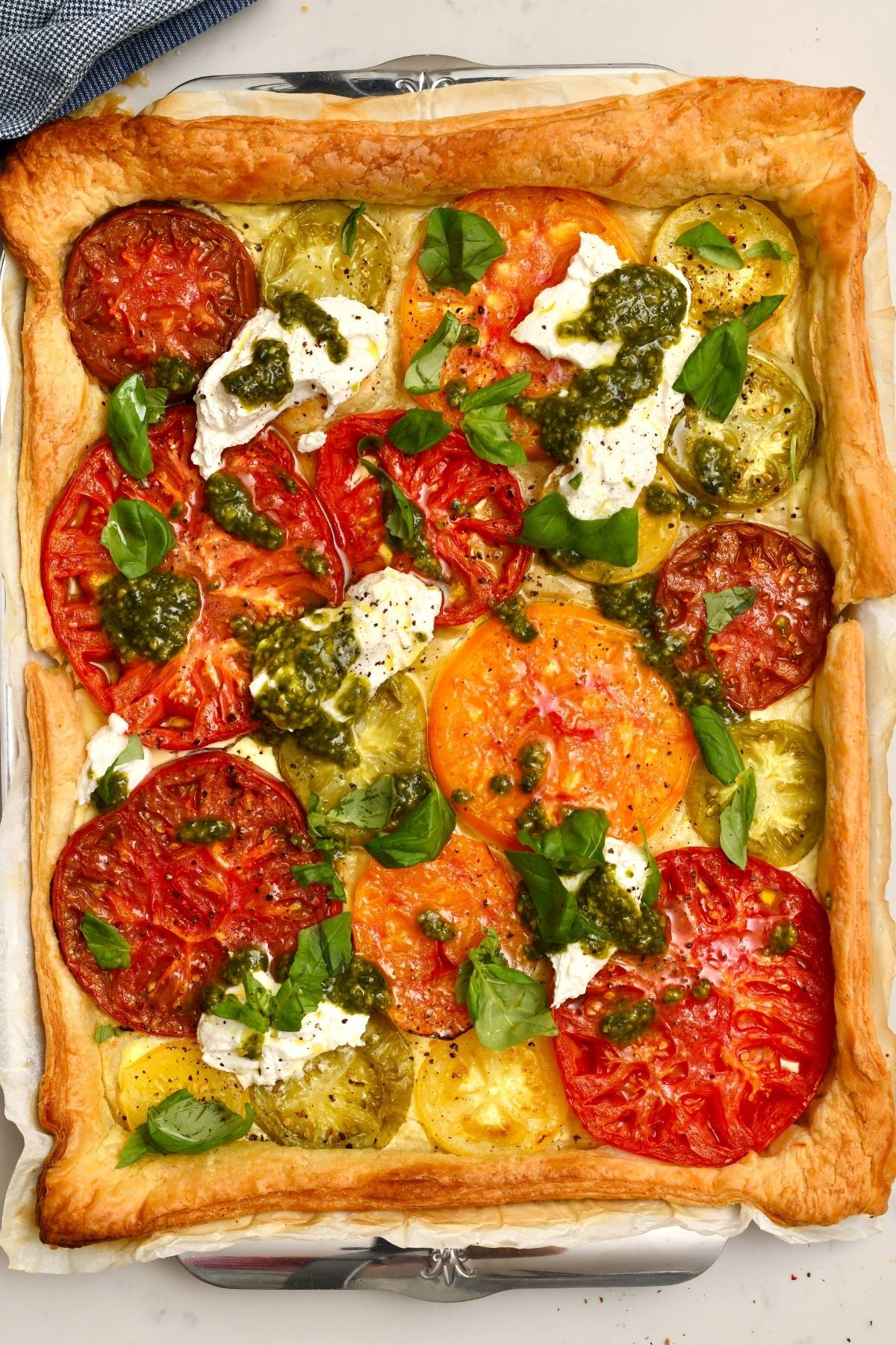 A top-down view of a freshly baked heirloom tomato tart with a golden puff pastry crust, topped with dollops of fresh cheese, spoonfuls of pesto, and fresh basil leaves.