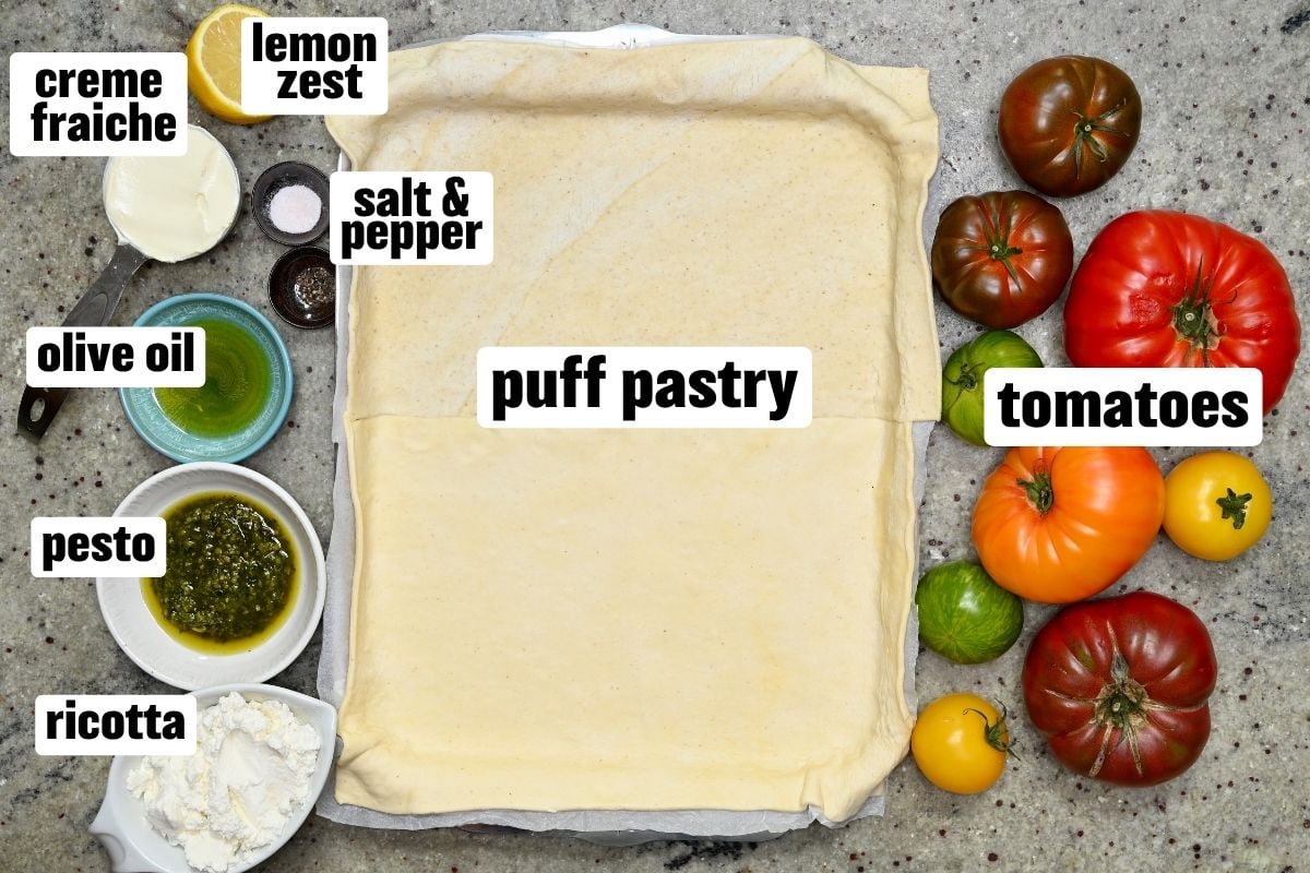 A top-down view of tomato tart ingredients surrounding a rolled-out sheet of puff pastry, including crème fraîche, lemon zest, salt and pepper, olive oil, pesto, ricotta, and colorful heirloom tomatoes.