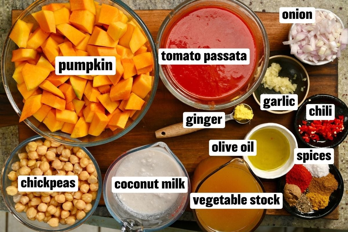 A flat-lay view of pumpkin curry ingredients, including diced pumpkin, chickpeas, tomato passata, coconut milk, and spices in bowls on a wooden board.